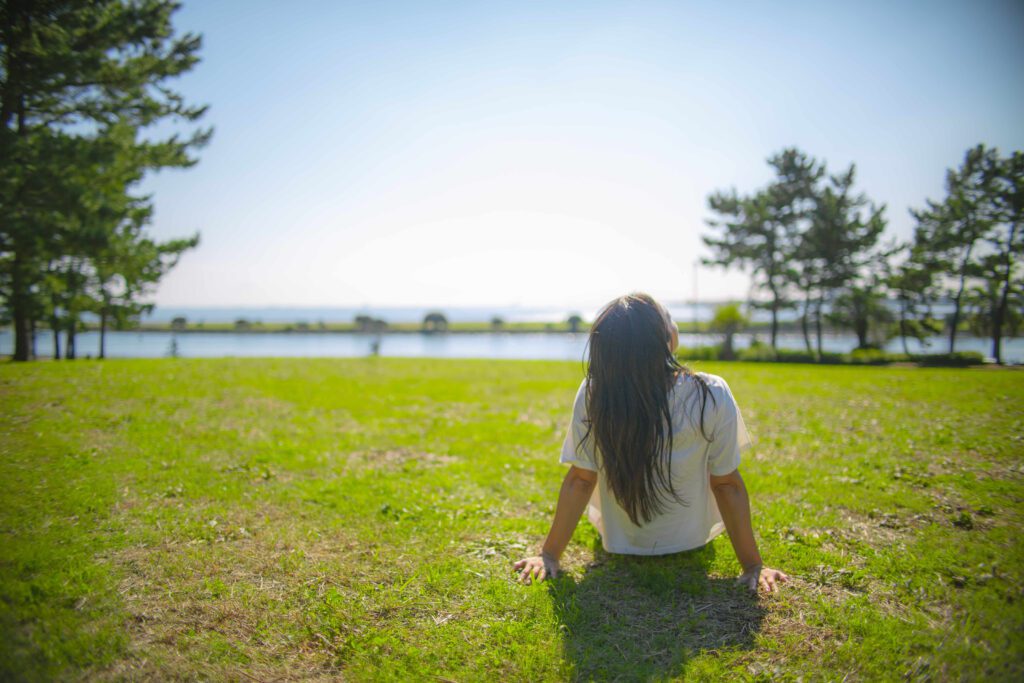 A woman sitting on grass looking out to the open with the sea in front of her meditating on the word of God and enjoying the presence of God. She's feeling peace and comfort through her practice of christian meditation and mindfulness. 